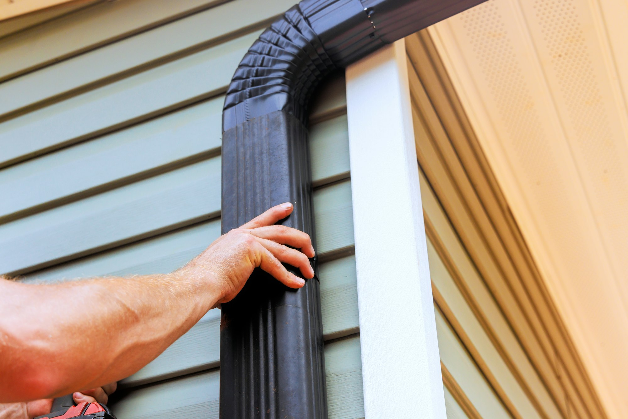 On roof, worker is installing rain gutter fitting a downpipe downspout bend